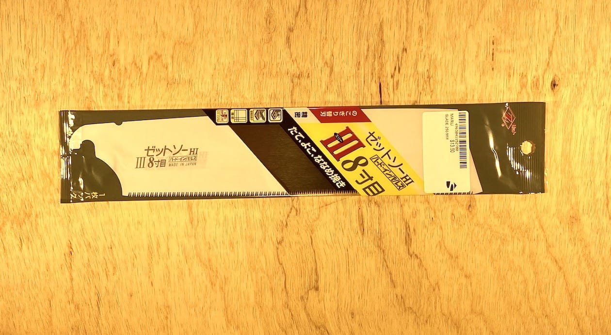 A package of the Universal Saw 250 III by Z-Saw, an 8-tooth thin saw blade from the MARU brand, is placed on a wooden surface. The packaging features a design in black, white, and yellow and contains Japanese text, making it ideal for fine woodworking projects.