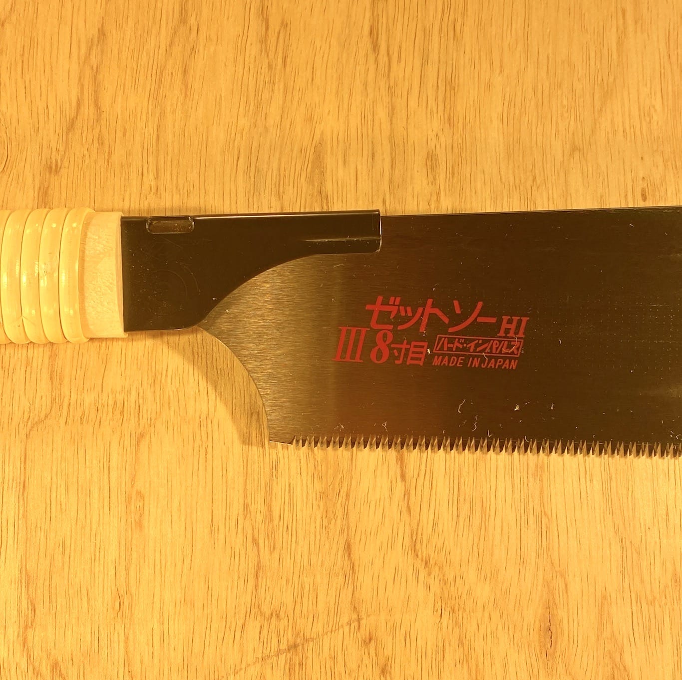 A Japanese handsaw, the Universal Saw 250 III by Z-Saw from the brand MARU, with a beige handle and red text on its thin blade reading "Z-Saw III 8" and "Made in Japan," lies on a wooden surface. Its universal teeth make it perfect for fine woodworking.