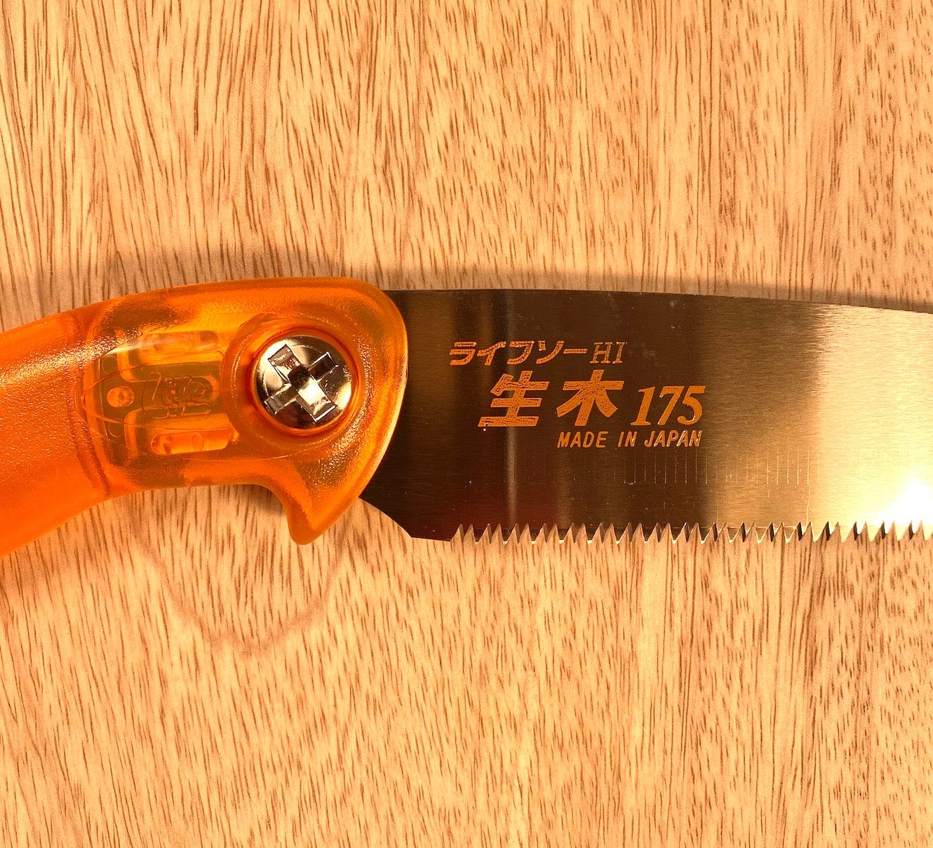 A close-up of a MARU Z-Saw Universal 175 Green Wood Pruner with an orange handle. The blade is engraved with Japanese characters, "HI 175," and "Made in Japan." Ideal for garden maintenance, it rests on a light wooden surface.
