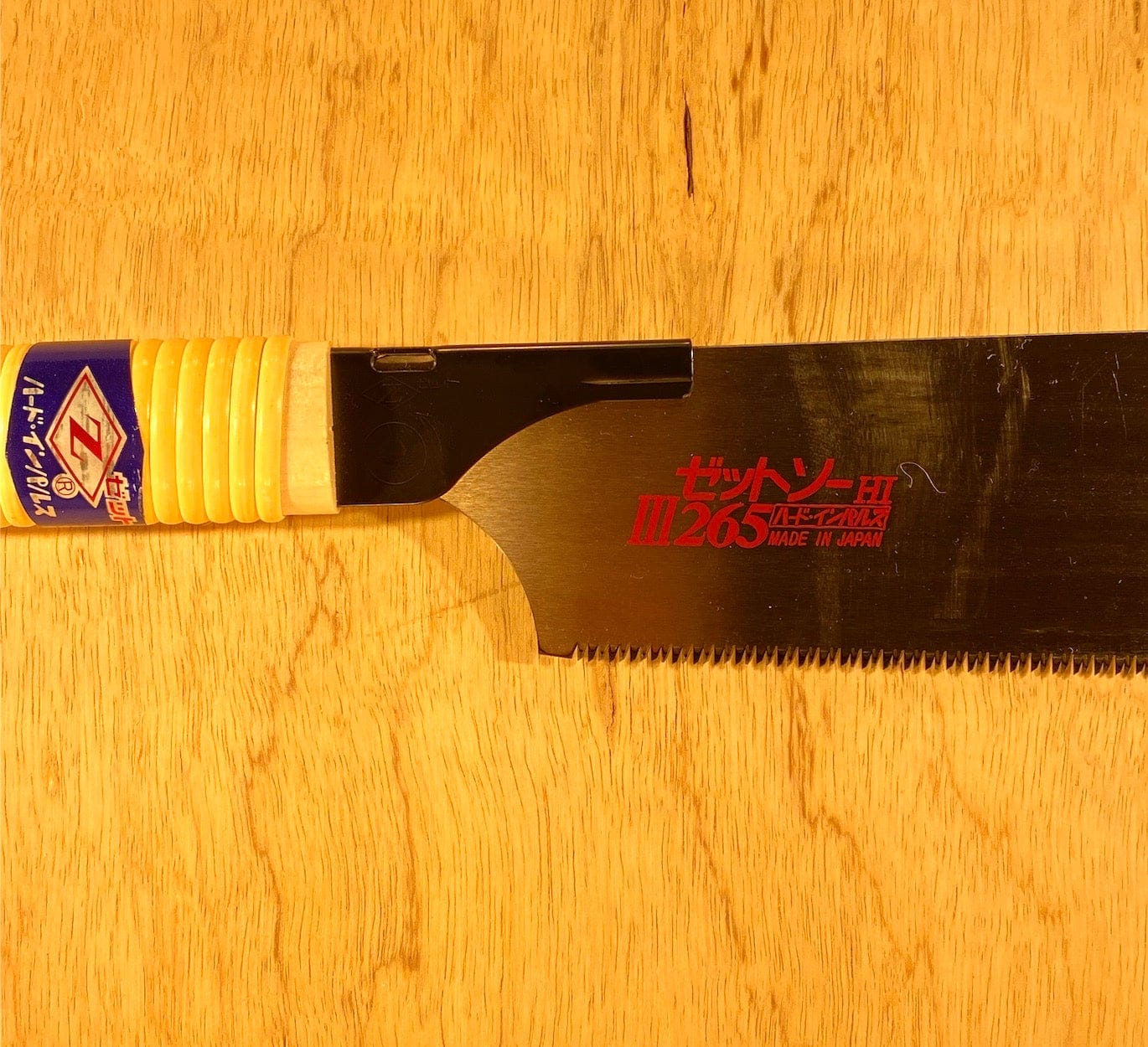 The Z-Saw Universal Saw 265, a versatile cutting tool by MARU with chisel-like teeth, features a yellow and blue handle labeled ZETSAW 265, resting elegantly on a wooden surface.