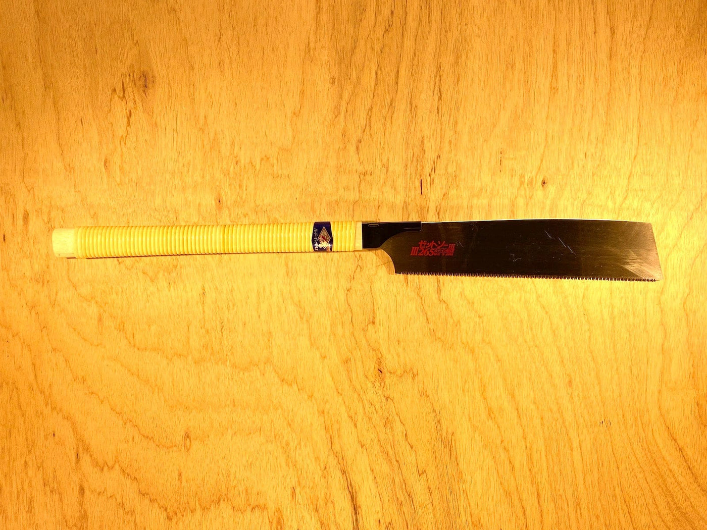 A Z-Saw Universal Saw 265 by MARU, featuring a yellow wrapped handle, rests on a wooden surface. It has a wide blade with chisel-like teeth and red branding near the handle.