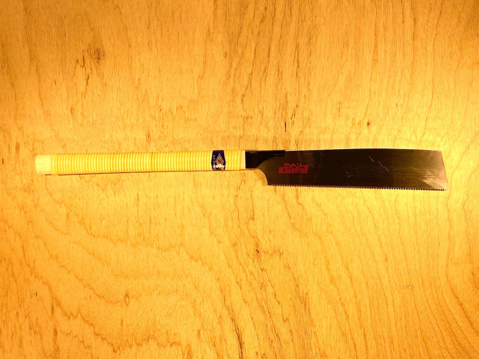 A Z-Saw Universal Saw 265 by MARU, featuring a yellow wrapped handle, rests on a wooden surface. It has a wide blade with chisel-like teeth and red branding near the handle.