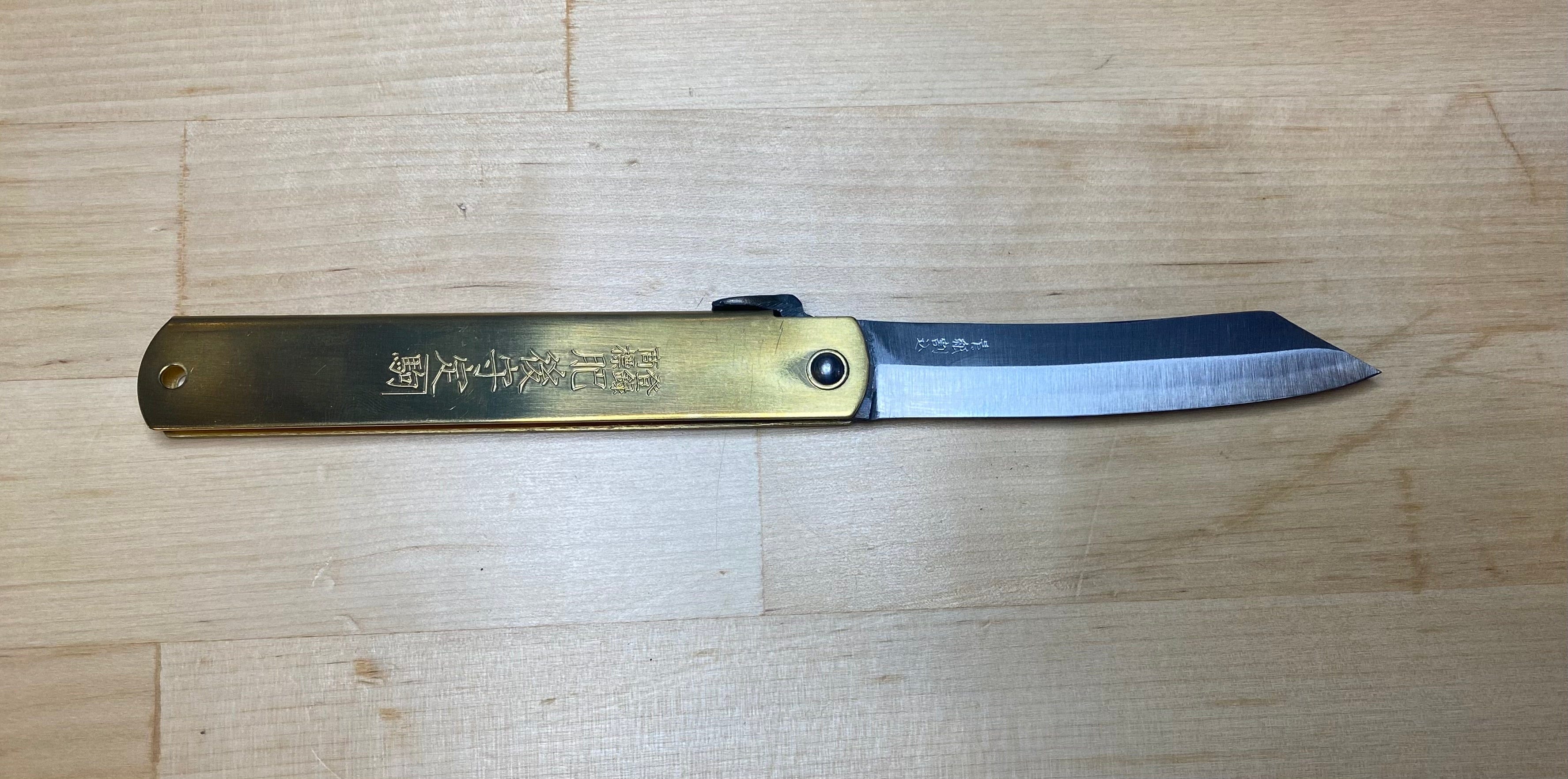 A MARU Higonokami Folding Knife Large Blue Steel Blade from Japan, featuring a brass handle with engraved characters, lies open on a light wooden surface.