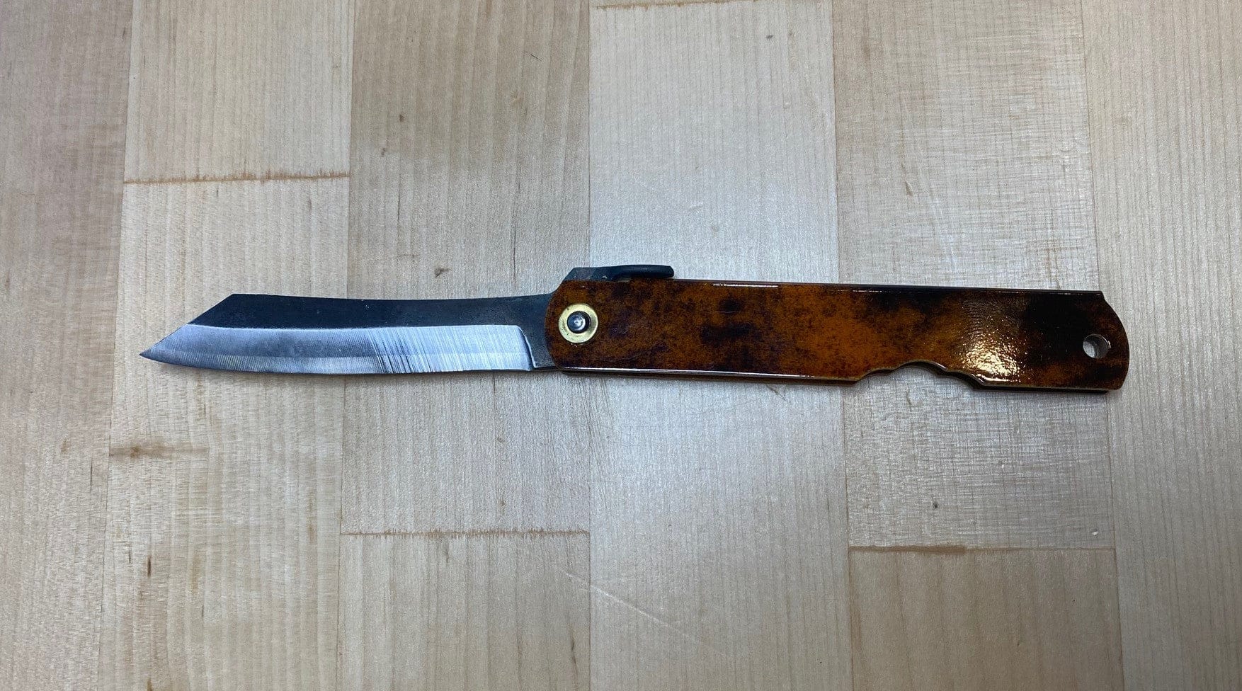 A MARU Higonokami Folding Knife Large Blue Steel Blade from Japan with a brown, rust-like handle and partially opened blade rests on a light wooden surface.