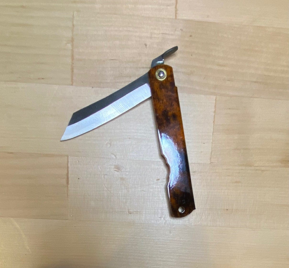 The MARU Higonokami Folding Knife Large Blue Steel Blade from Japan, featuring a patterned brown handle and shiny partially open blade, rests on a light wood surface.