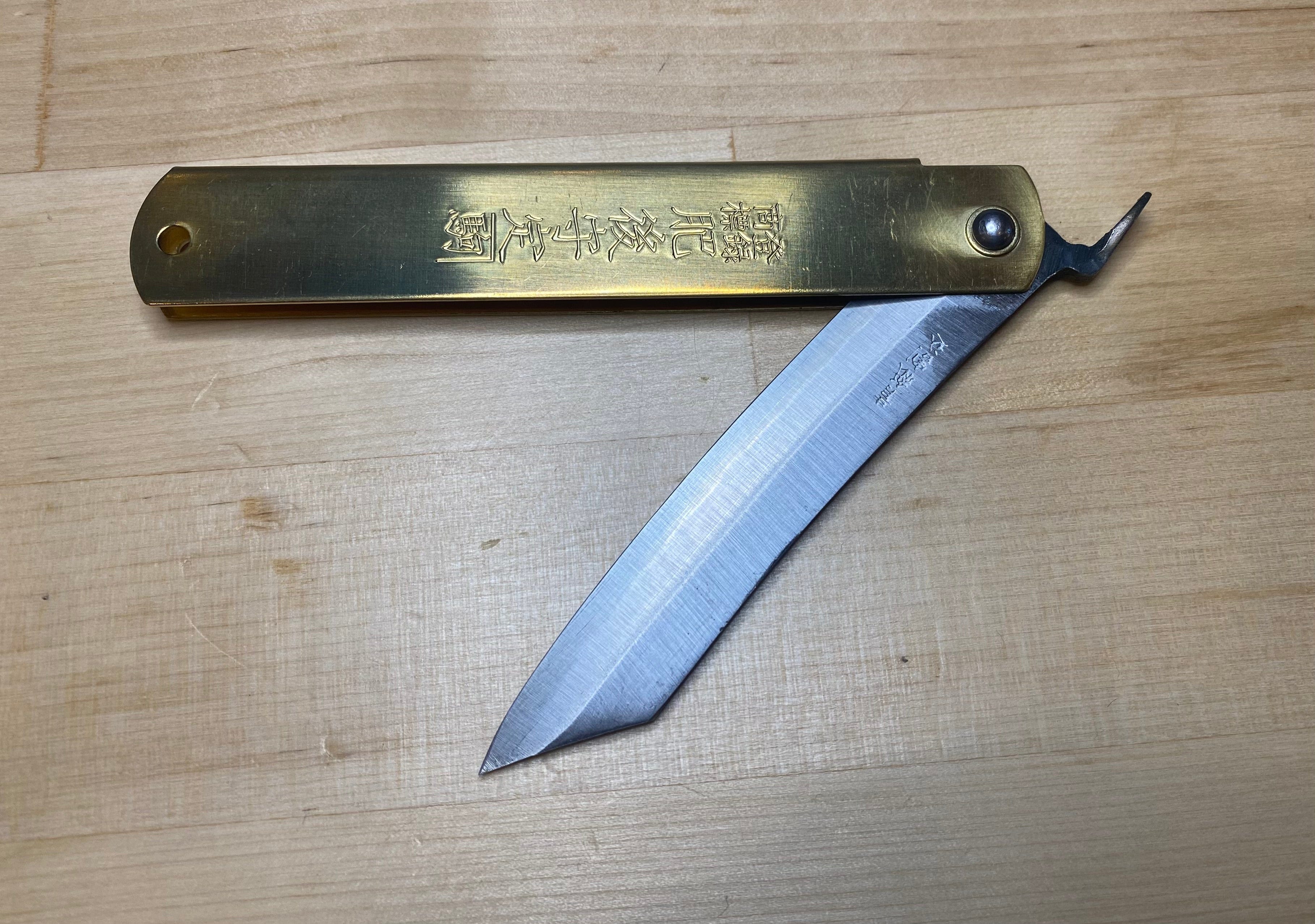 The MARU Higonokami Folding Knife Large Blue Steel Blade from Japan, featuring a brass handle engraved with Japanese characters, rests on a wooden surface with its blade partially open.