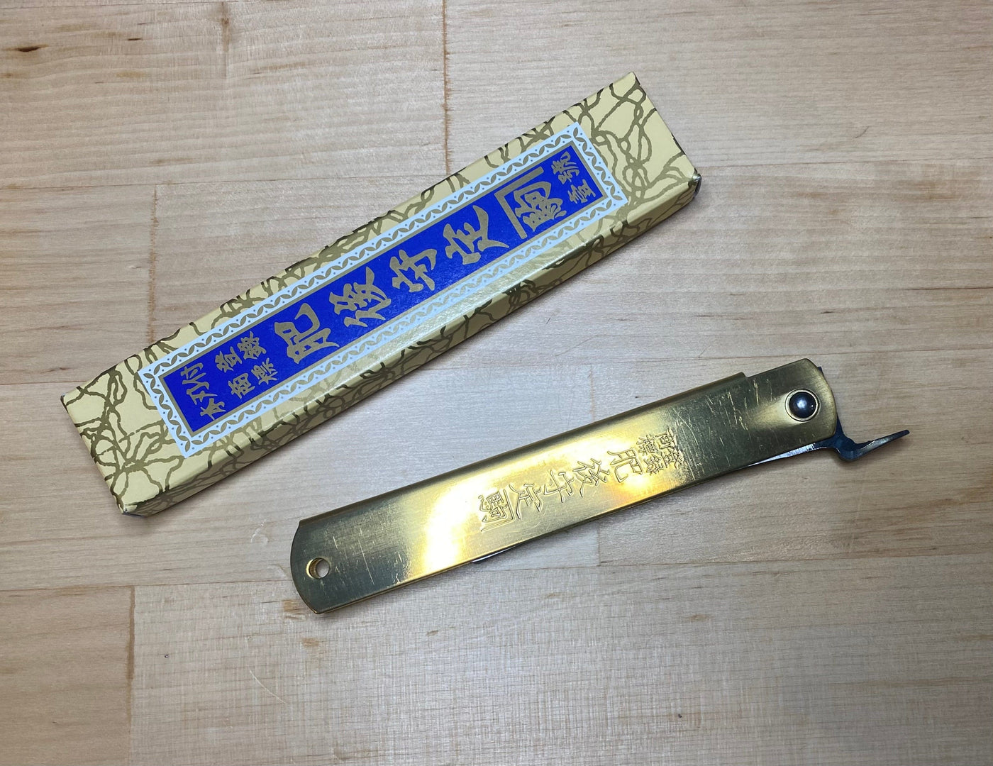 The MARU Higonokami Folding Knife Large Blue Steel Blade from Japan with a brass handle and engraved characters rests beside its yellow and blue box on a light wooden surface.