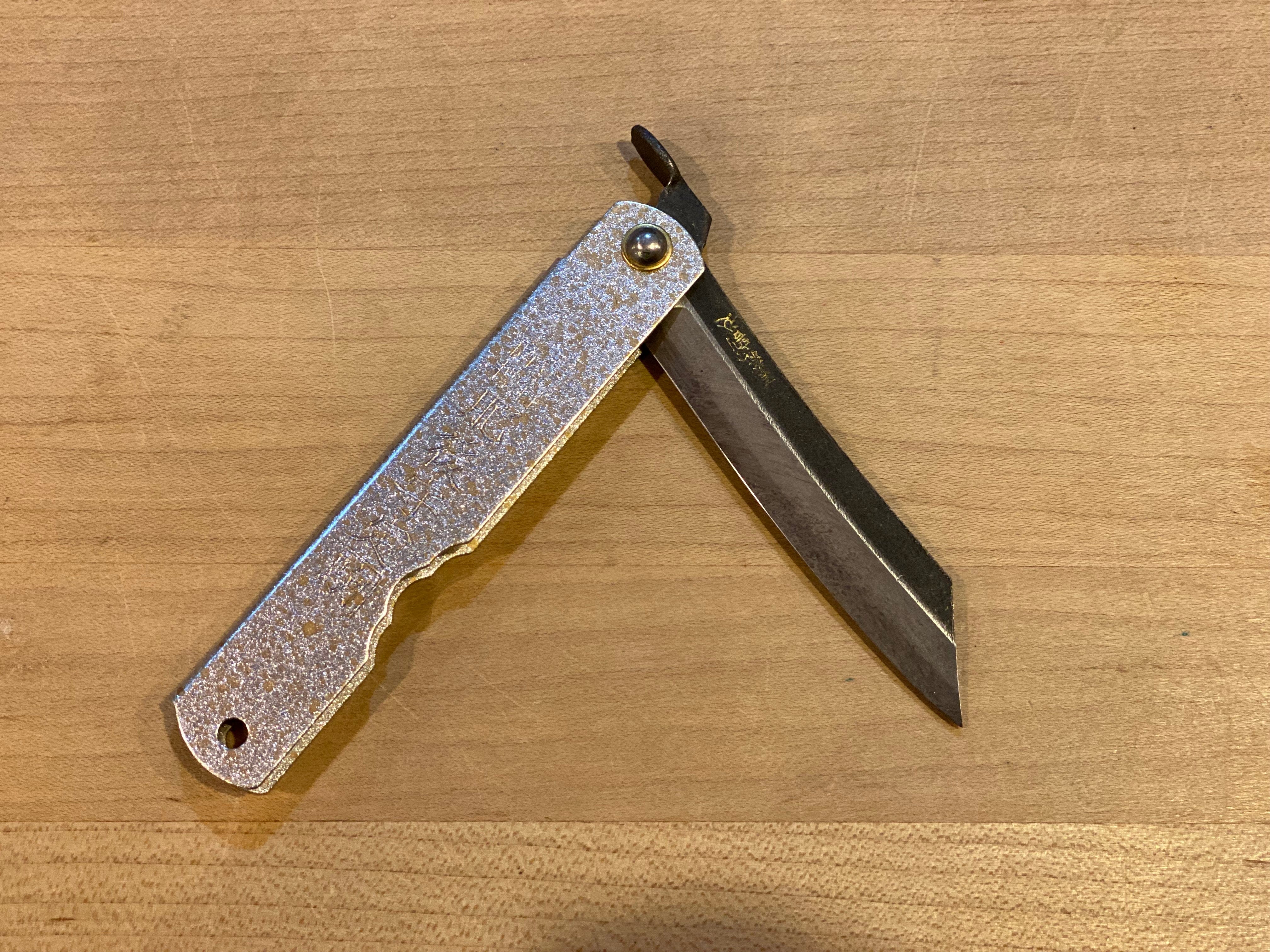 The MARU KNIFE FOLDING AOGAMI STEEL with a silver and gold handle is shown partially open, displaying its straight-edged blade on a wooden surface.