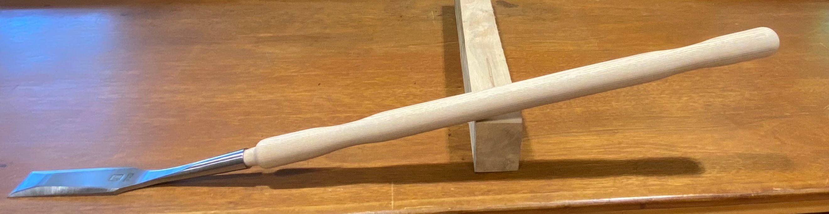 The MHG Timber Framing Slick, a woodworking tool made from high-alloy material, sits with its wooden handle on a stand atop a polished surface.