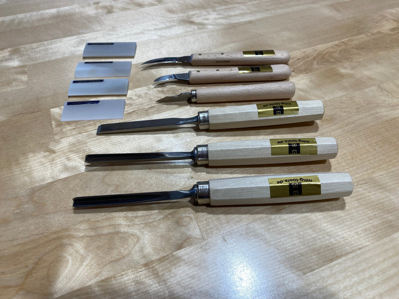 The MHG Carving Kit includes four chisels and three flat-edged blades with wooden handles and metal blades, neatly arranged on a light wooden surface—ideal for use with an Arkansas oil whetstone.