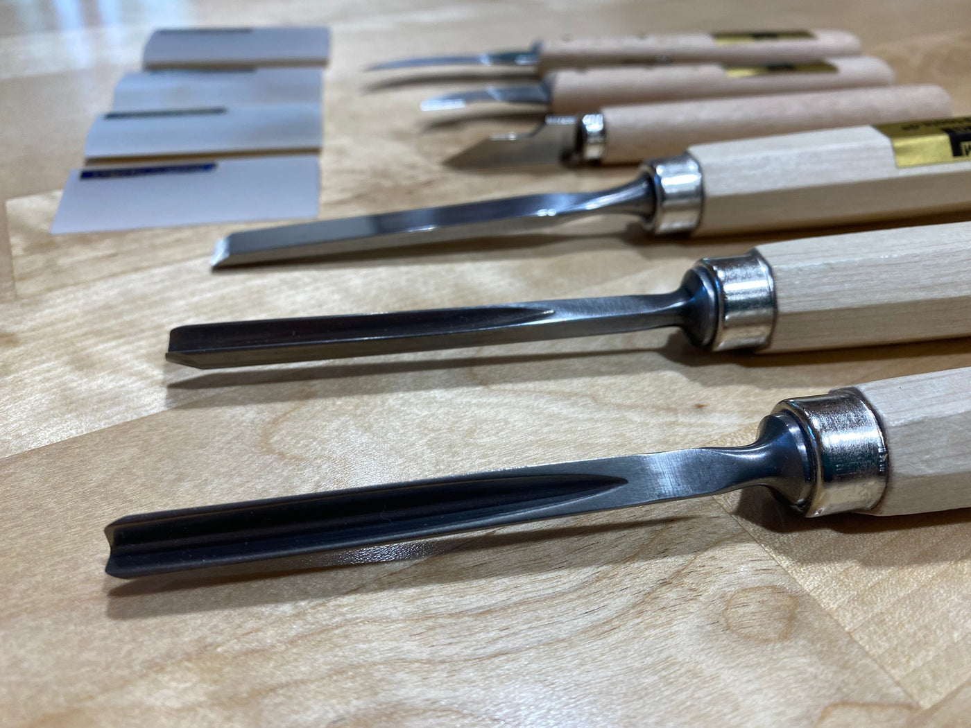 A close-up of the MHG Carving Kit shows woodworker tools with wooden handles neatly arranged on a light wooden surface, featuring small flat tools and a sharpening stone in the background.