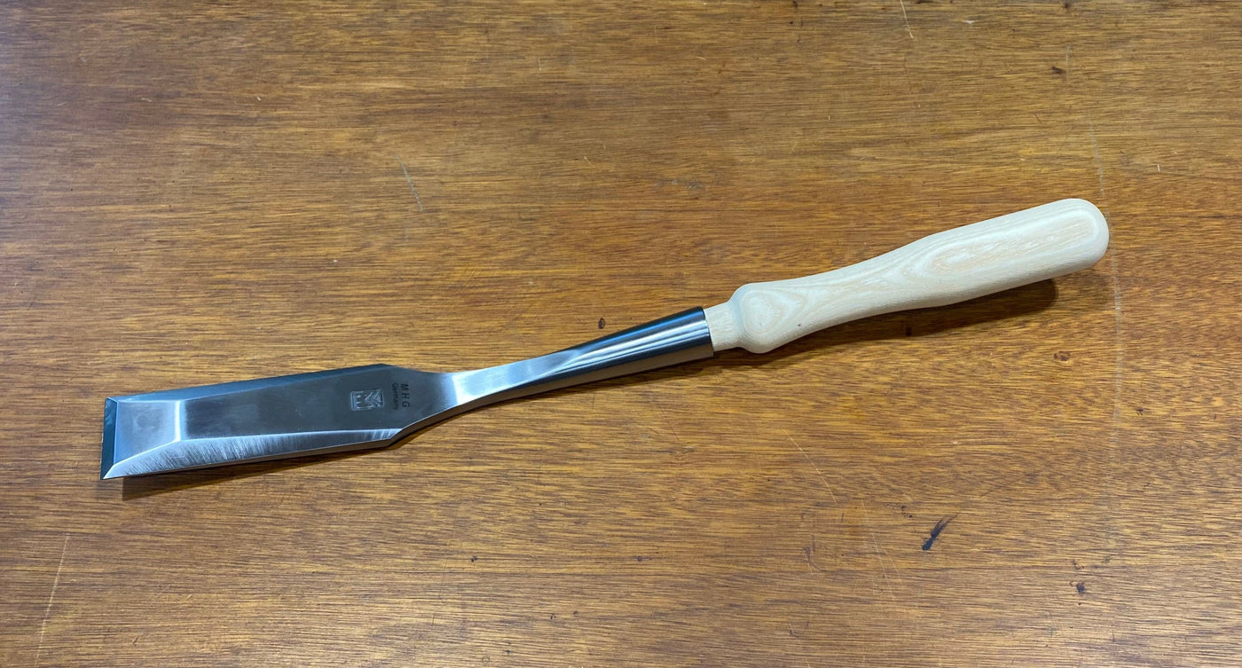 The MHG Timber Framing Slick, crafted with a light wooden handle and a polished high-alloy metal blade, elegantly rests on a wooden surface, embodying the precision necessary for expert woodworking.