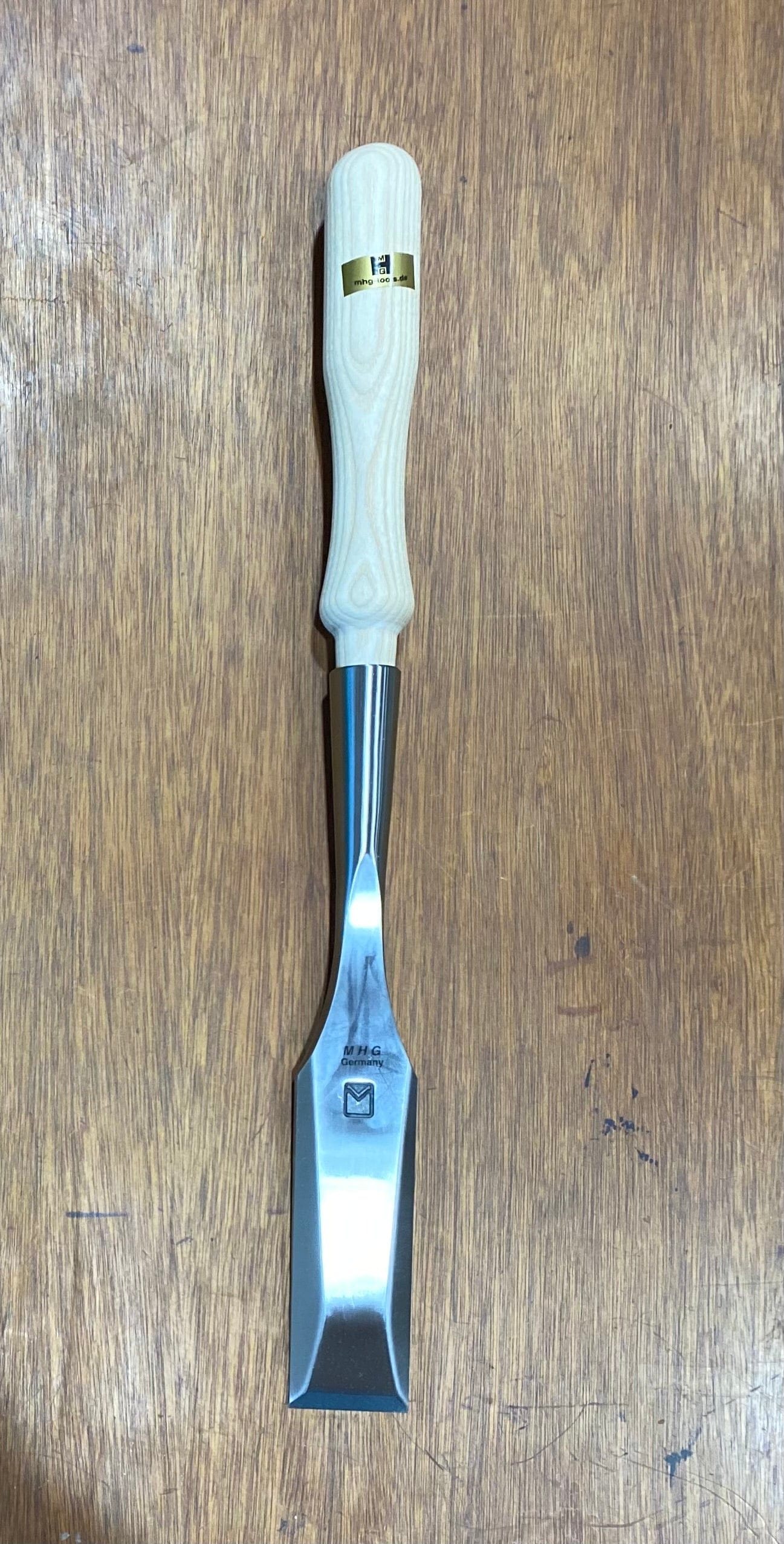 The MHG Timber Framing Slick, featuring a smooth wooden handle and a gleaming metal blade, rests elegantly on a wooden surface, highlighting the craftsmanship of this essential woodworking tool.