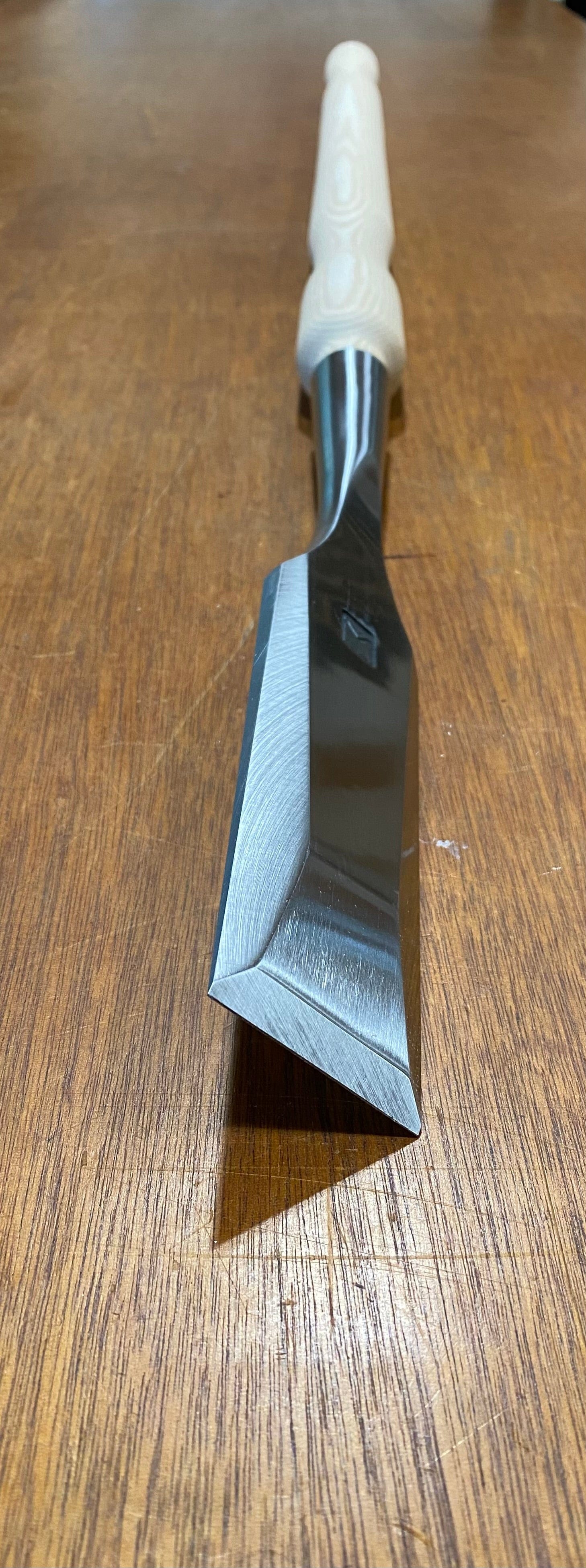 The MHG Timber Framing Slick, a woodworking tool with a wooden handle and a sharp metal blade made from high-alloy material, is pictured resting on a wooden surface.