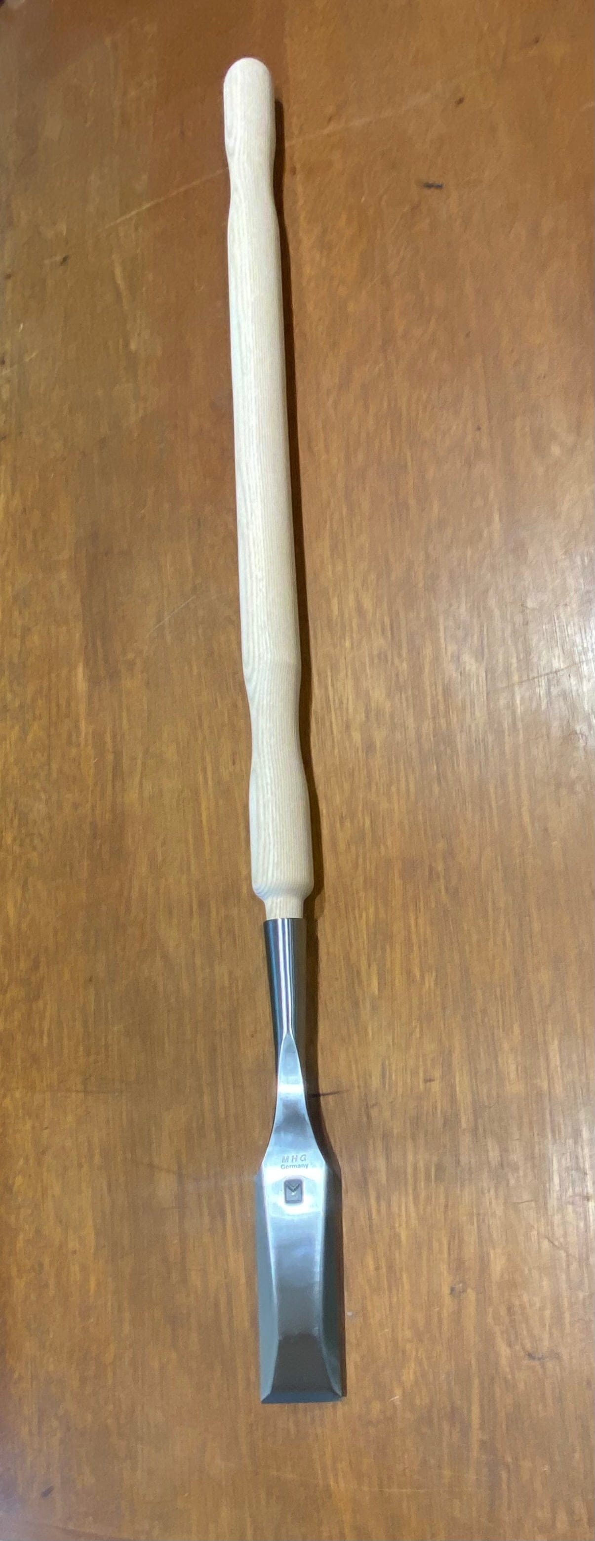 An MHG Timber Framing Slick featuring a wooden handle and a high-alloy metal blade, placed on a wooden surface.