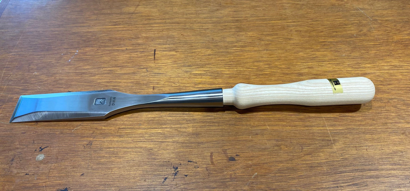A woodworking tool, the MHG Timber Framing Slick features a wood handle and a polished, high-alloy metal blade, elegantly resting on a wooden surface to embody precision.