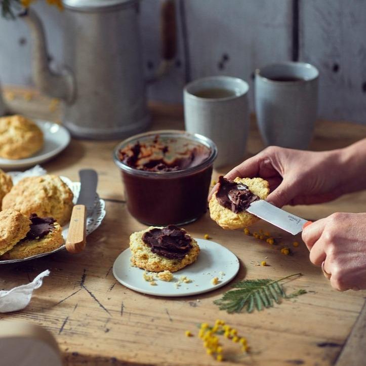 Hands spreading chocolate on a biscuit with the Opinel Brunch Spreading Knife. A jar of spread, more biscuits, mugs, and a metal watering can are in the background on a wooden table, perfect for a cozy picnic.