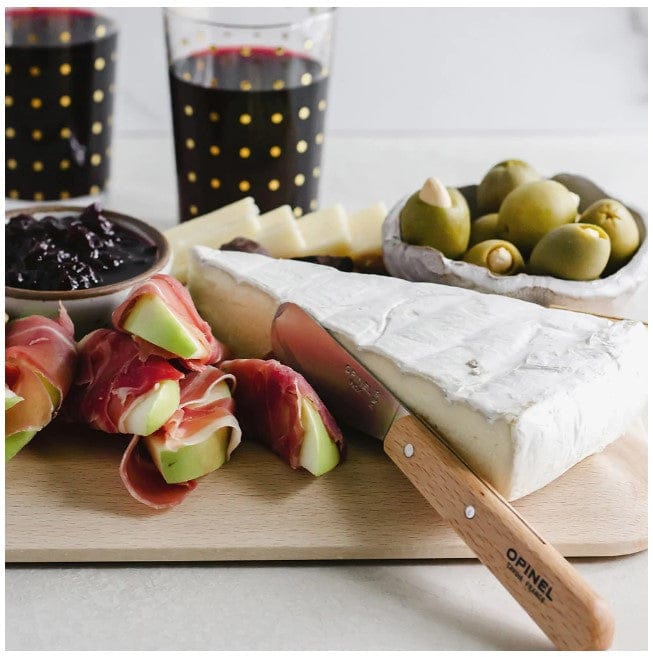 A classic cheese board with brie, apple slices in prosciutto, dark jam, green olives, and two red wine glasses. An Opinel Essential Spreading Knife with a wooden handle and high-carbon stainless steel blade rests elegantly on the board.
