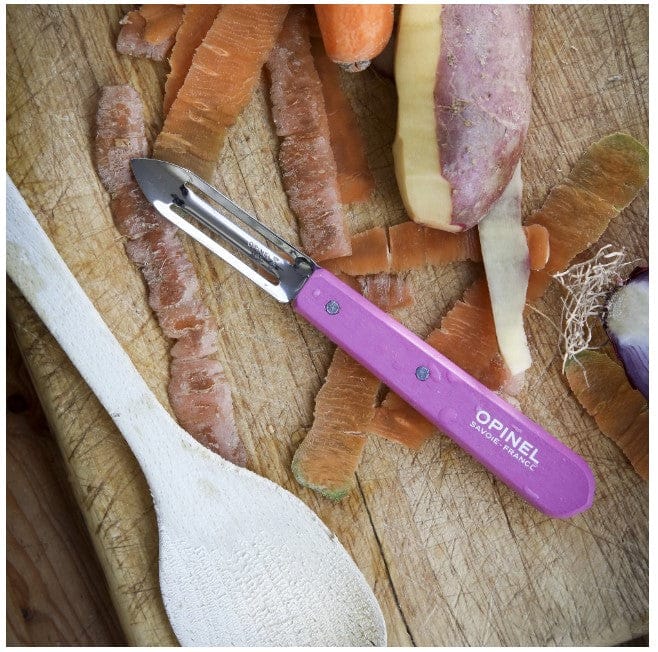 An Essential Stationary Peeler by Opinel, featuring a stainless steel blade and purple handle, sits on a wooden cutting board with potato skins, a potato, carrot, and onion piece. A reliable kitchen tool companion, the wooden spoon complements this vibrant cooking scene.