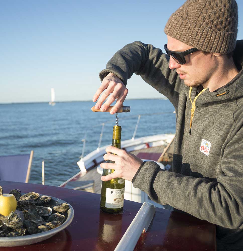 Person in a beanie and sunglasses opens a wine bottle with an Opinel professional-quality stainless steel corkscrew from the KNIFE OPINEL CORKSCREW AND CHEESE KNIFE set next to a plate of oysters on a boat.