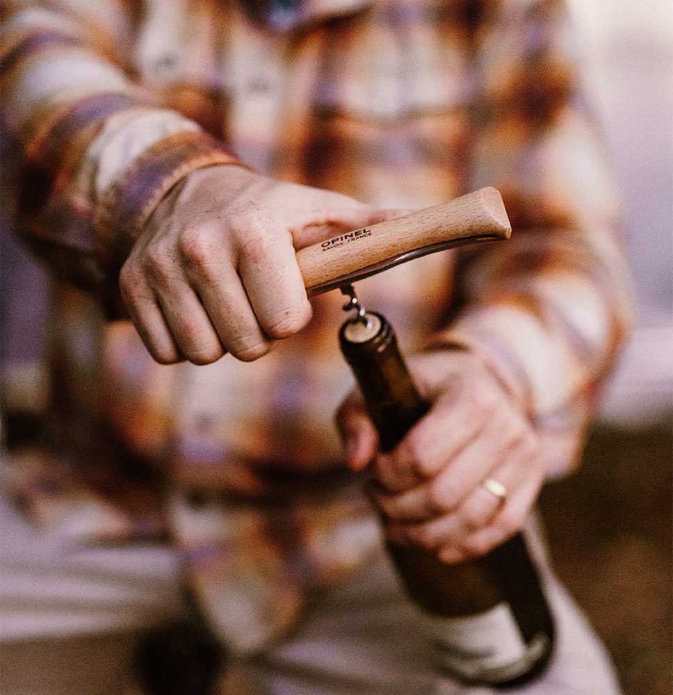 A person in a plaid shirt is using the professional-quality Opinel KNIFE OPINEL CORKSCREW AND CHEESE KNIFE to open a wine bottle.