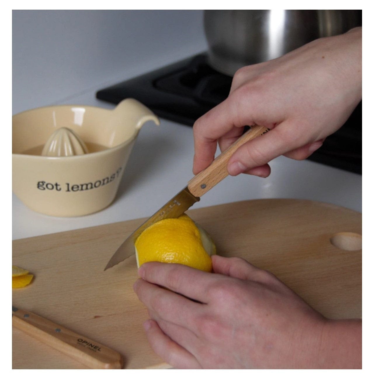 A person slices a lemon with the Opinel Small Kitchen Essentials from Shelter Institute on a wooden cutting board near a cream-colored got lemons? juicer, with a stove and pot in the background—showcasing must-have kitchen tools.