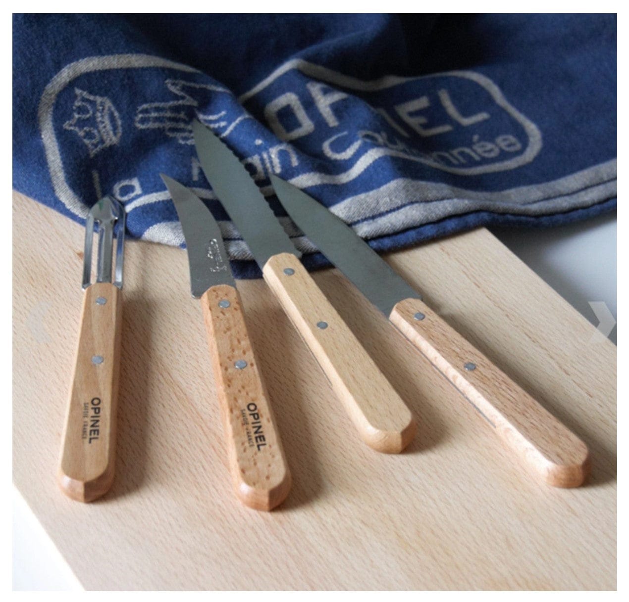 The Shelter Institute Opinel Small Kitchen Essentials set includes three versatile knives and a peeler with wooden handles, displayed on a wooden cutting board and accompanied by a blue Opinel cloth.