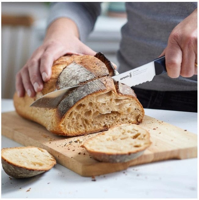 A person slices rustic bread on a wooden board using the Opinel Bread Knife OPINEL USA Intempora 8 with a sharp serrated blade. Two bread slices and scattered crumbs rest nearby.