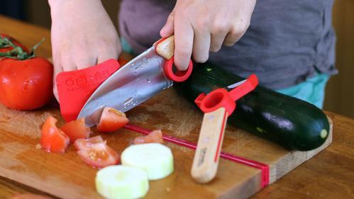 A person slices vegetables, including tomatoes and cucumbers, on a wooden cutting board while wearing finger guards on both hands, using the Opinel Le Petit Chef kitchen knife with stainless steel blades.