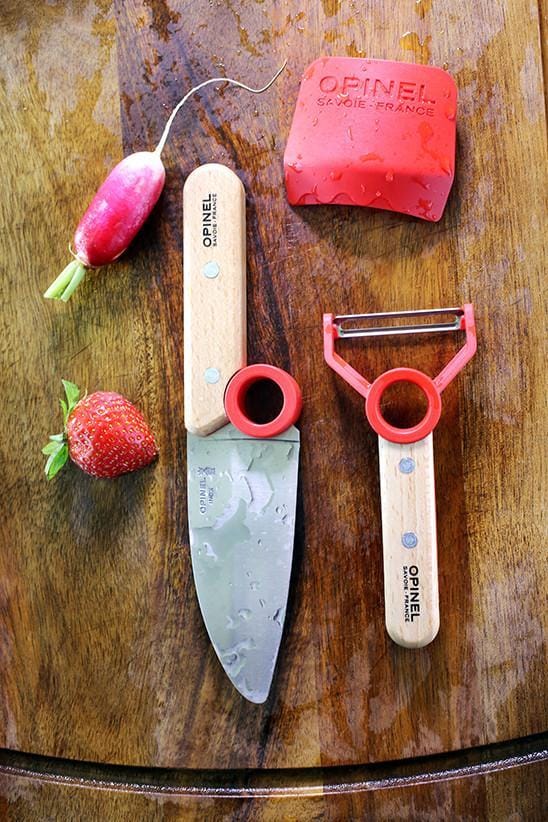 The Le Petit Chef by Opinel features a wooden cutting board that holds a knife with stainless steel blades, a peeler, a sliced strawberry, and a radish. The knife and peeler are designed with wooden handles adorned with red accents, promoting children cooking safety while serving as educational kitchen tools.