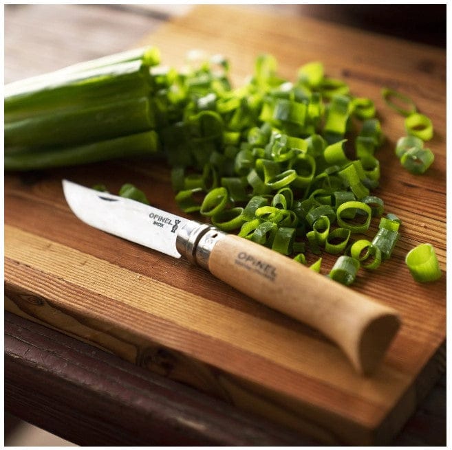 An Opinel No.08 Folding Knife with a wooden handle and carbon steel blade rests on a cutting board next to sliced green onions.
