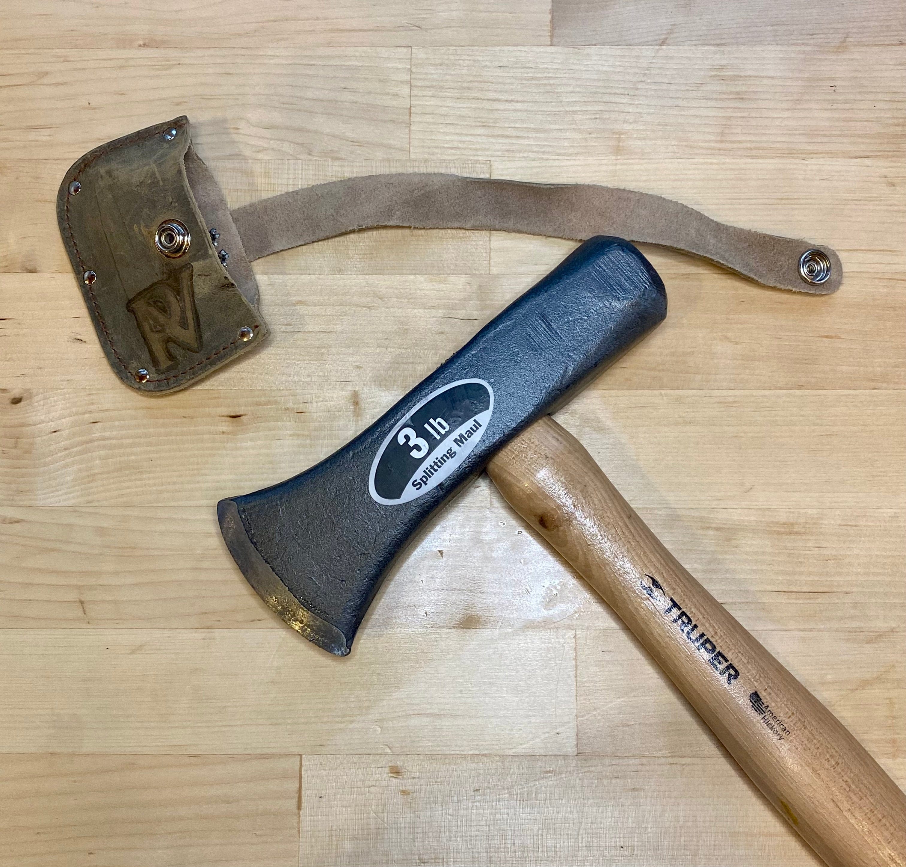 A 3 lb Peavey Mini Splitting Maul by PEA with a wooden handle rests on wood next to a detached leather blade guard with snap closure—ideal for outdoor activities and camping gear collections.