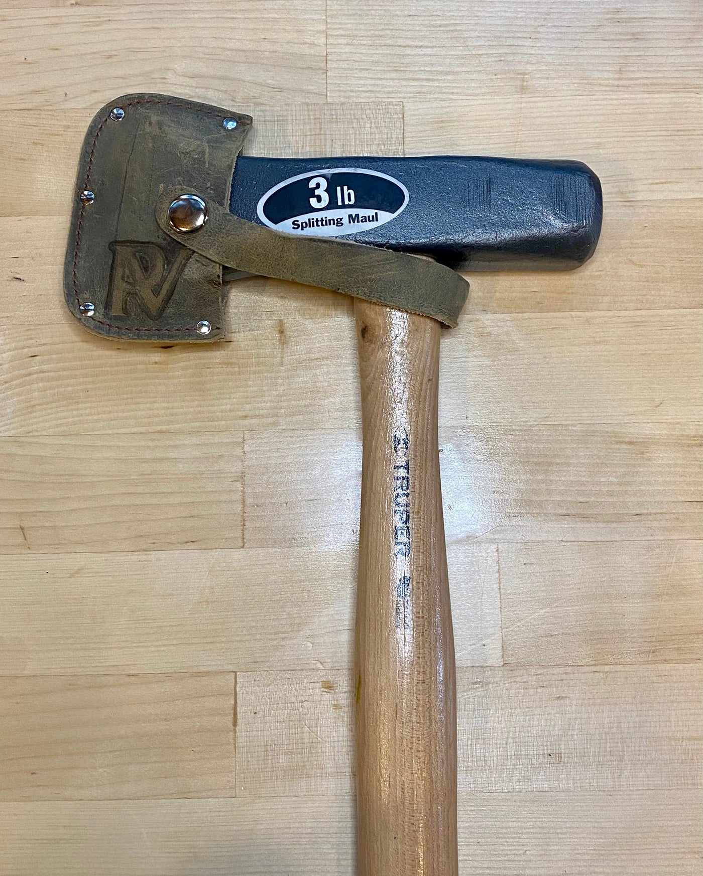 The PEA Peavey Mini Splitting Maul features a 3-lb black metal head, wooden handle, and leather blade guard—ideal for outdoor use or as essential camping gear, shown resting on a light wood surface.