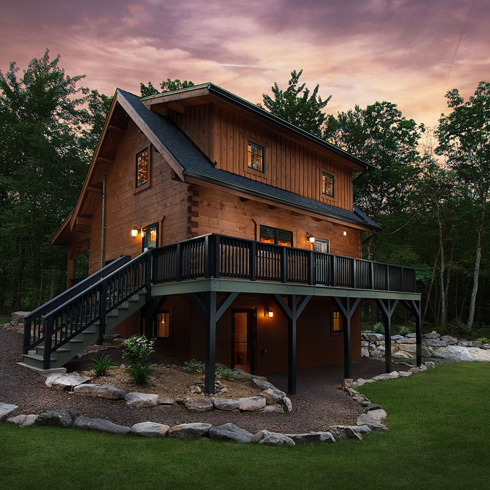 The two-story wooden cabin with a wraparound porch, nestled among trees and illuminated by exterior lights at dusk, showcases the exceptional wood preservation achieved with OUT's Q8 LOG OIL.