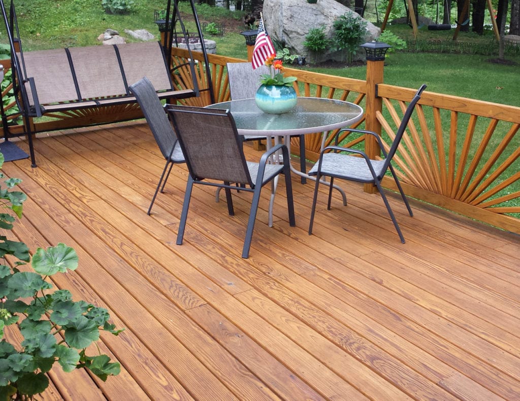 A beautiful wooden deck featuring a round glass table, four chairs, a swing, and a small American flag in a vase of flowers. Enhanced by the lush greenery in the background, this space radiates brightness and charm thanks to OUT's Q8 LOG OIL exterior wood preservation, which accentuates its natural beauty.