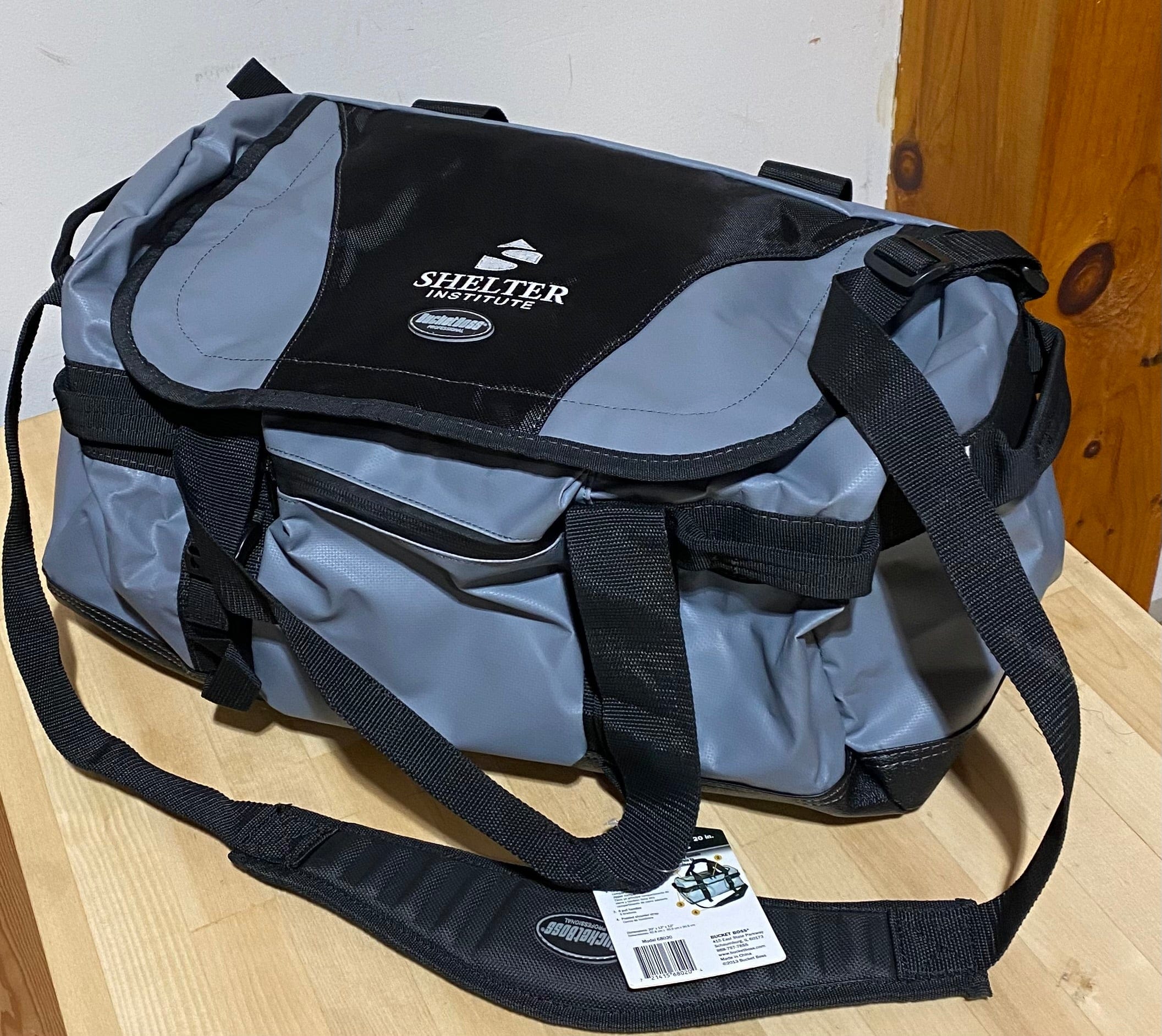 The Shelter Institute Duffel by PUL, a gray and black all-weather bag made from waterproof tarpaulin, has handles and pockets. It's placed on a wooden surface with a tag attached to one handle.