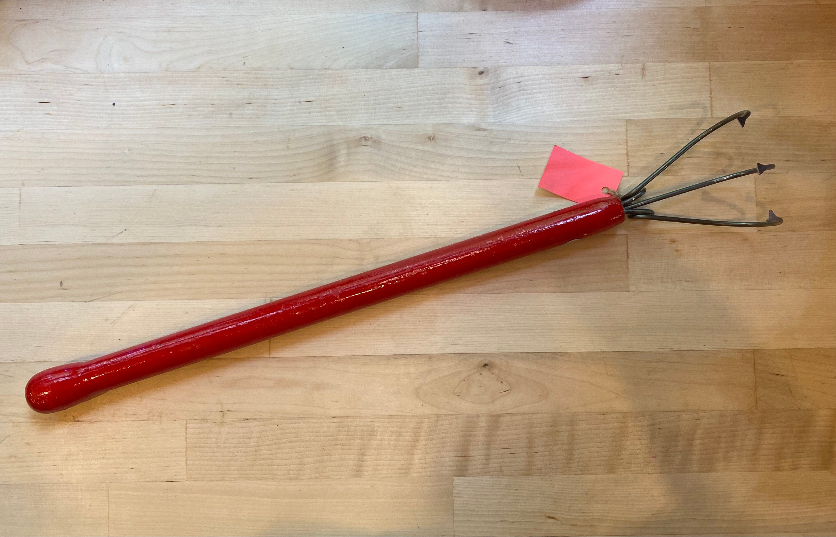 The Magic Weeder Gardening Tool by QMT, featuring a red metal handle and a three-pronged claw design, rests on a wooden surface—an ideal weeding solution for any gardener.