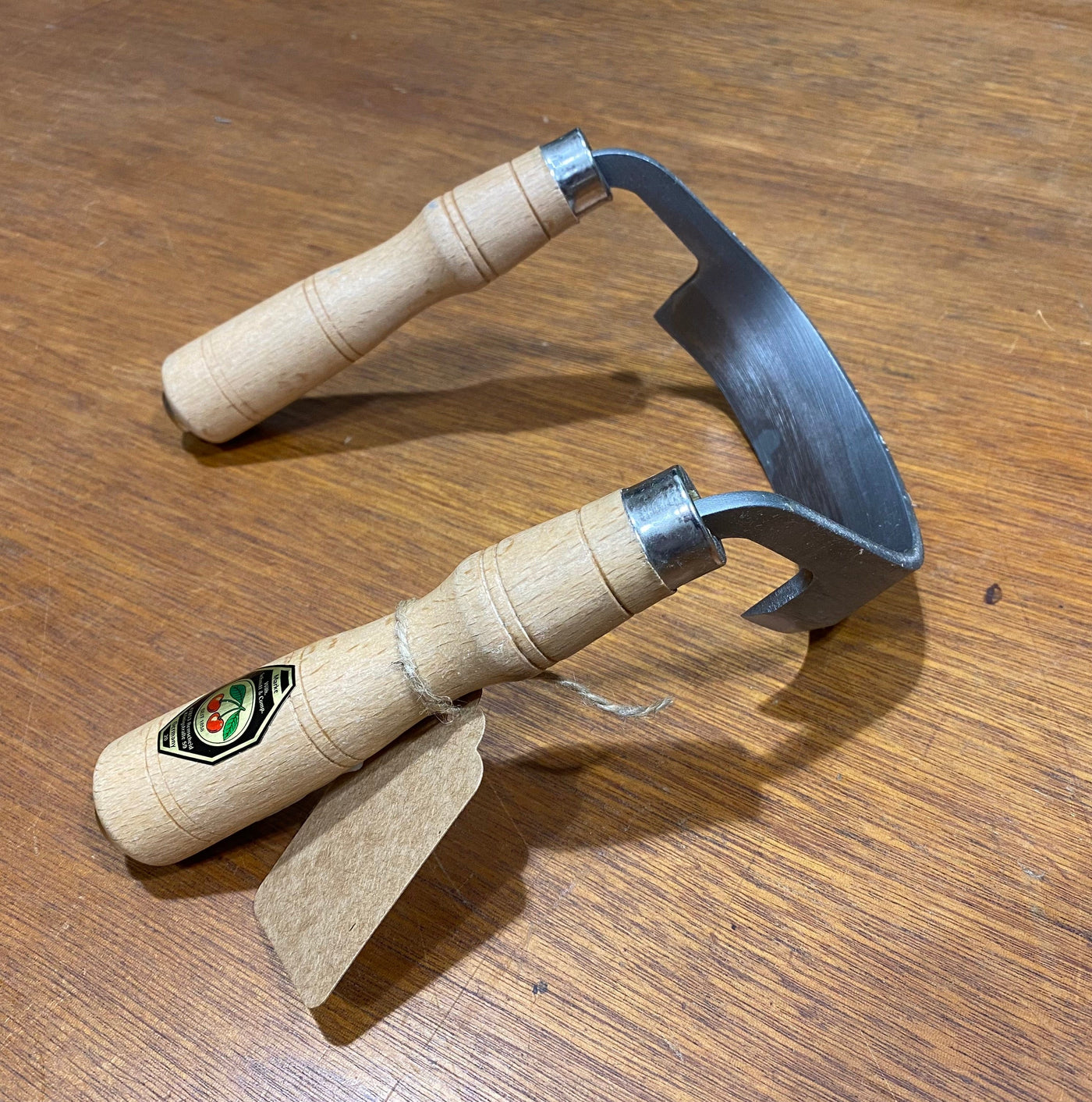 A ROB Two Cherries Inshave featuring two wooden handles, a curved metal blade, and a label rests on wood. One handle has a tagged string—perfect for bowl carvers and precise wood shaping.
