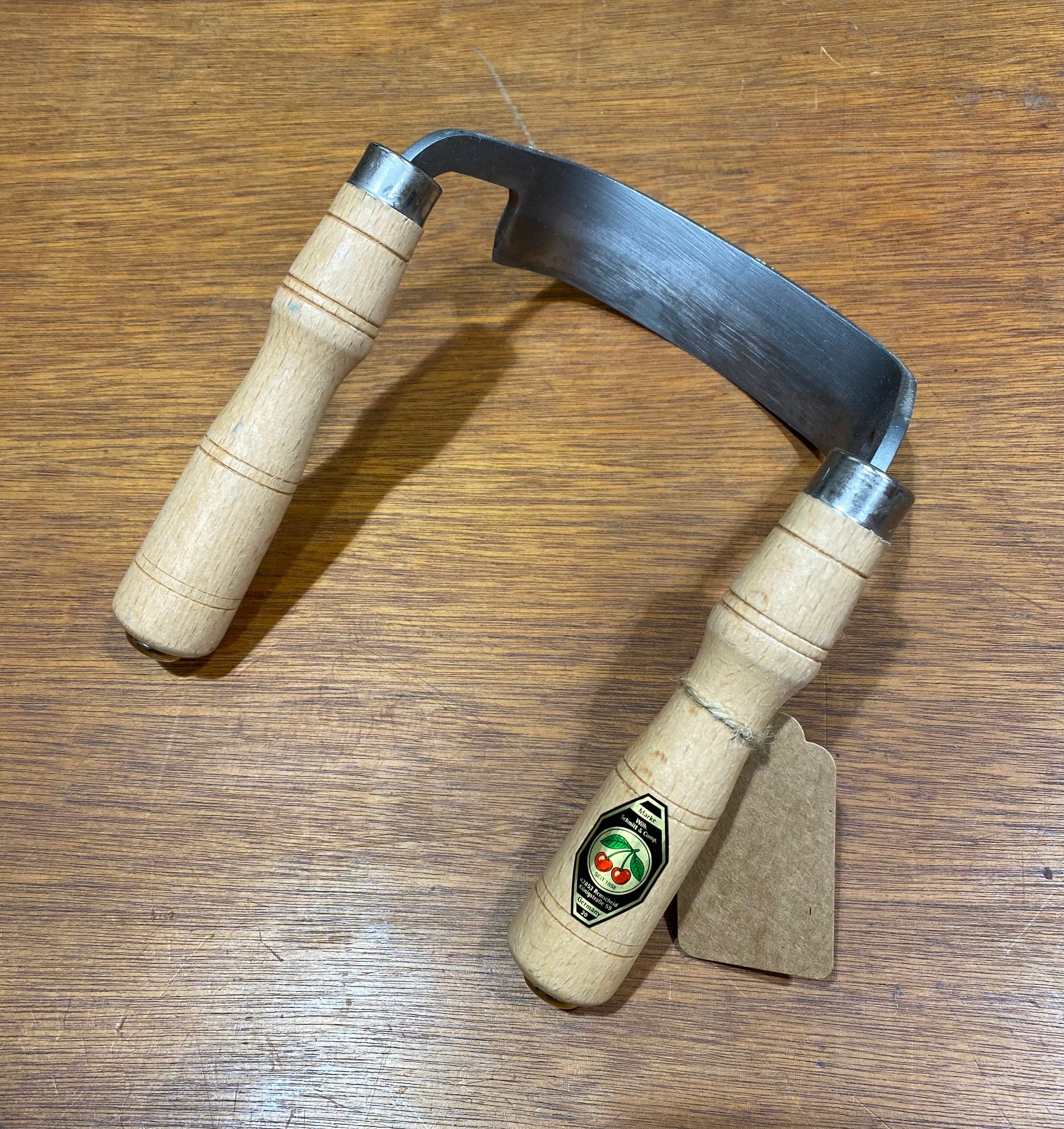 A ROB Two Cherries Inshave, a woodworking tool for bowl carving, rests on wood. It has wooden handles, a curved metal blade, a handle with a logo label, and an attached brown tag.
