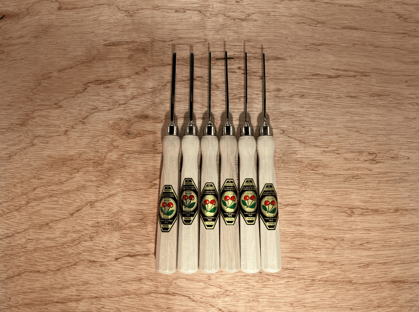 The ROB Two Cherries Micro Carving Tool Set of 6 features octagonal hornbeam handles with green and gold labels, black metal blades, and is lined up vertically on a wooden surface—an ideal set for micro carving.