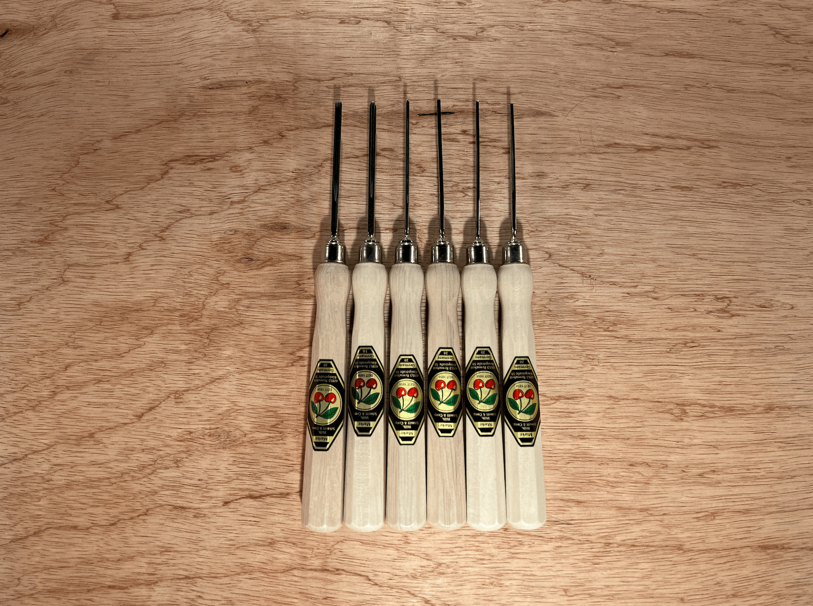 The ROB Two Cherries Micro Carving Tool Set of 6 features octagonal hornbeam handles with green and gold labels, black metal blades, and is lined up vertically on a wooden surface—an ideal set for micro carving.