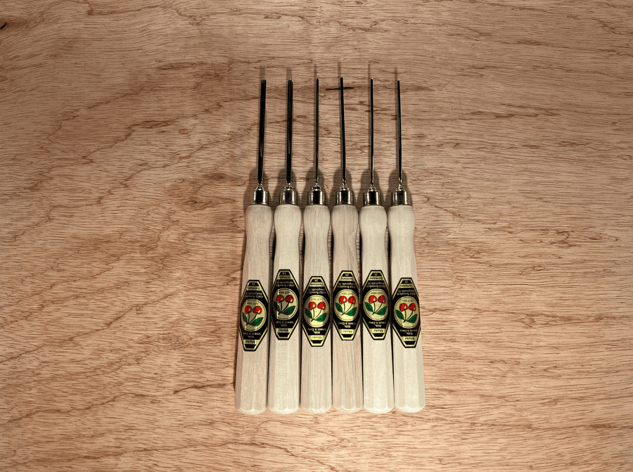The ROB Two Cherries Micro Carving Tool Set of 6 features octagonal hornbeam handles with green and gold labels, black metal blades, and is lined up vertically on a wooden surface—an ideal set for micro carving.