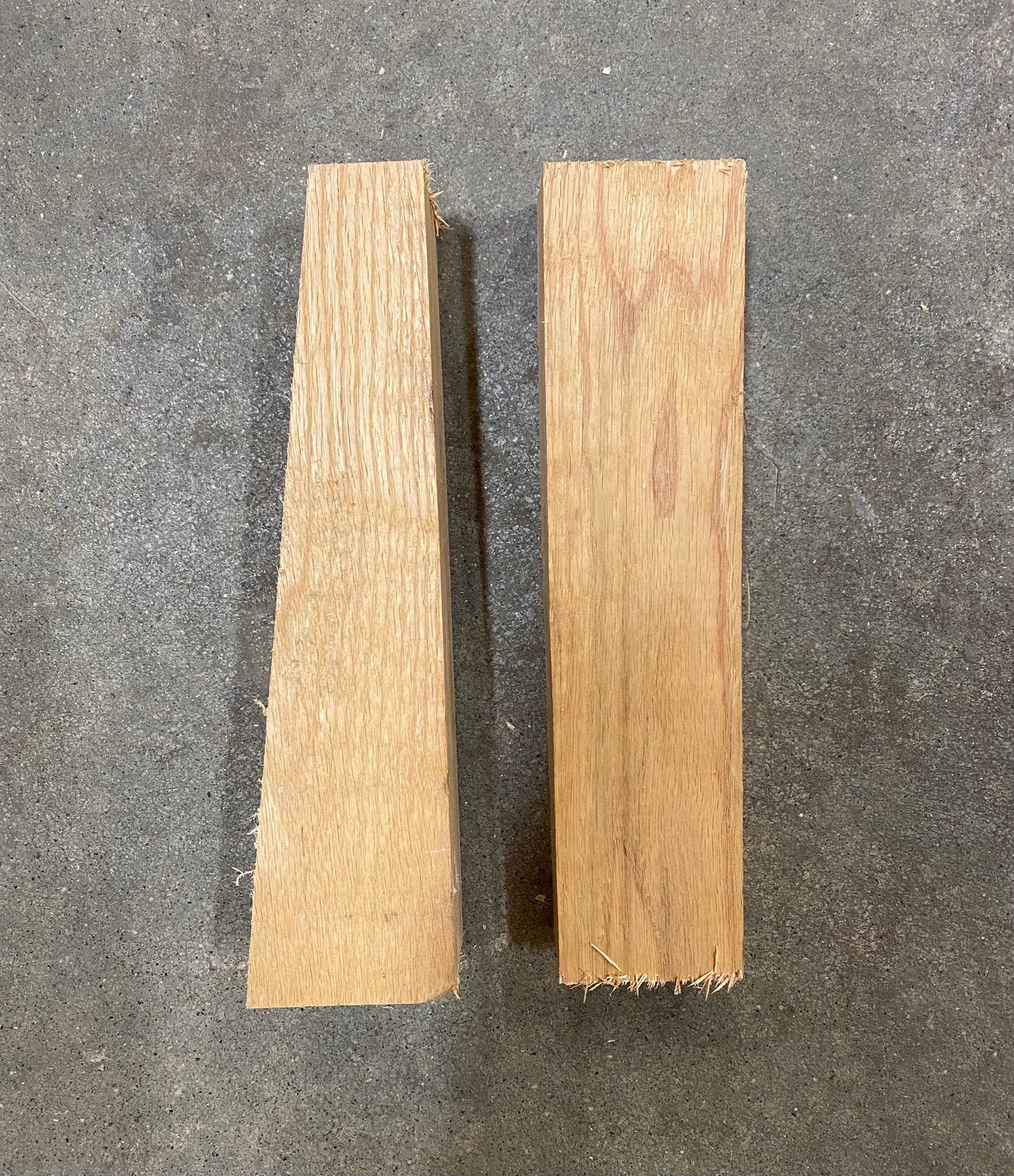 Two pieces of cut Red Oak from the Shelter Institute are standing vertically on a grey concrete floor. One piece is an Oak Wedge for Timber Frame, tapering from bottom to top, while the other piece is rectangular.