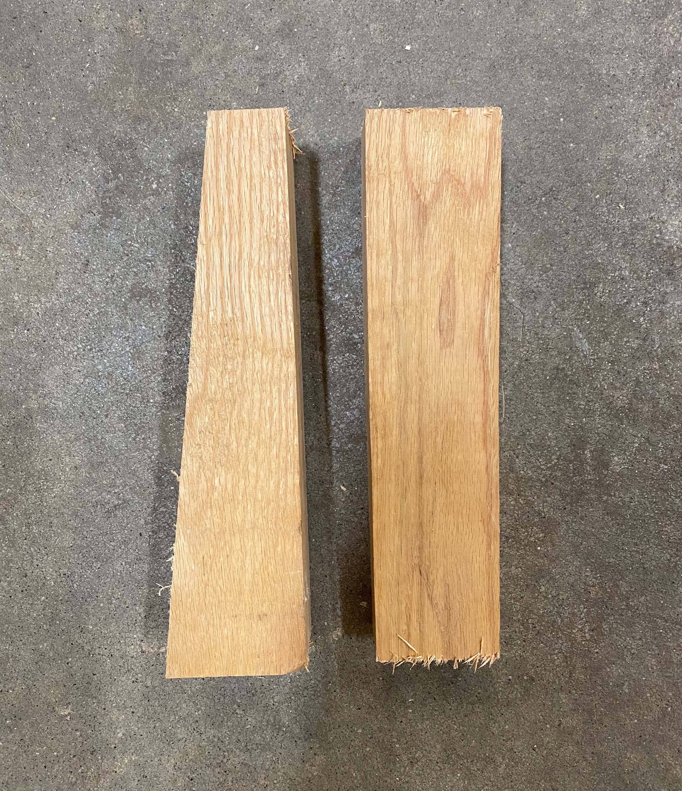 Two pieces of cut Red Oak from the Shelter Institute are standing vertically on a grey concrete floor. One piece is an Oak Wedge for Timber Frame, tapering from bottom to top, while the other piece is rectangular.