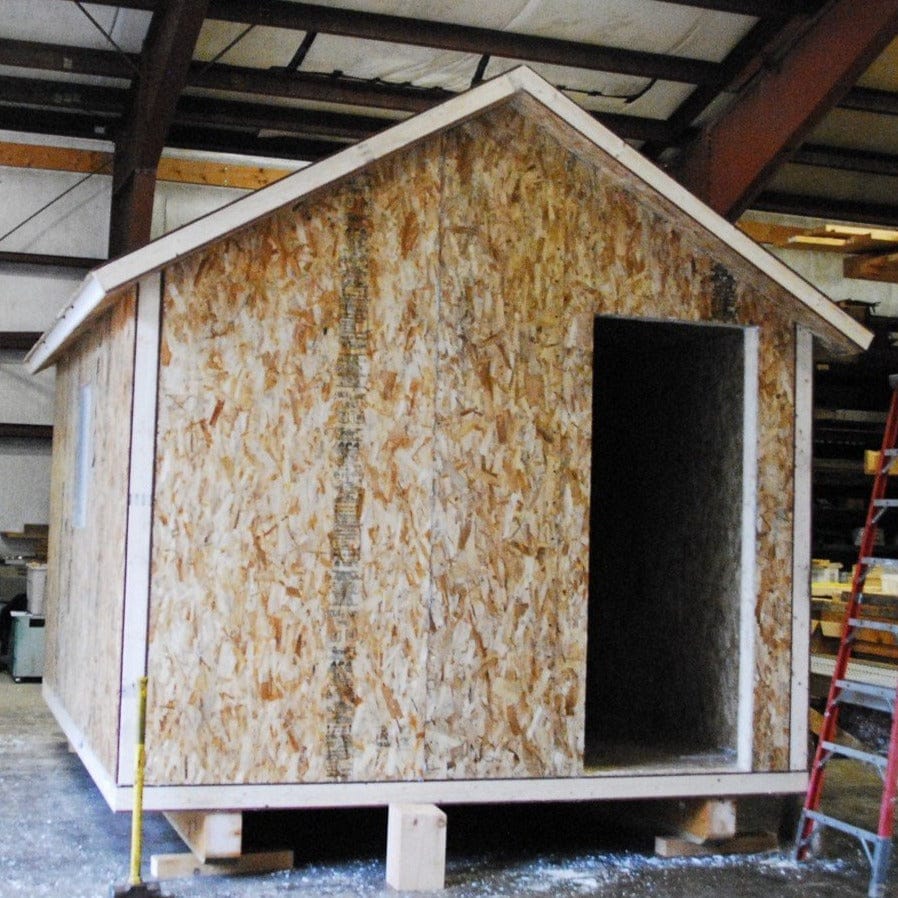 A 10x12 SIP Cottage by Shelter Institute, featuring unfinished oriented strand board walls and an open doorway, sits on wooden blocks inside a warehouse. A red ladder appears on the right.