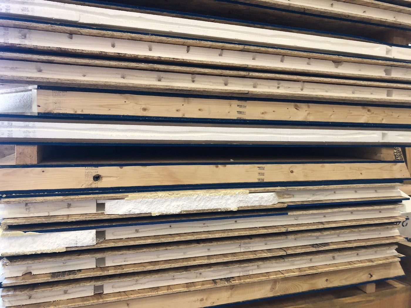 A pile of Shelter Institute's 10x12 SIP Cottage panels are neatly stacked in the workshop, showcasing the layers of wood and white insulating material.