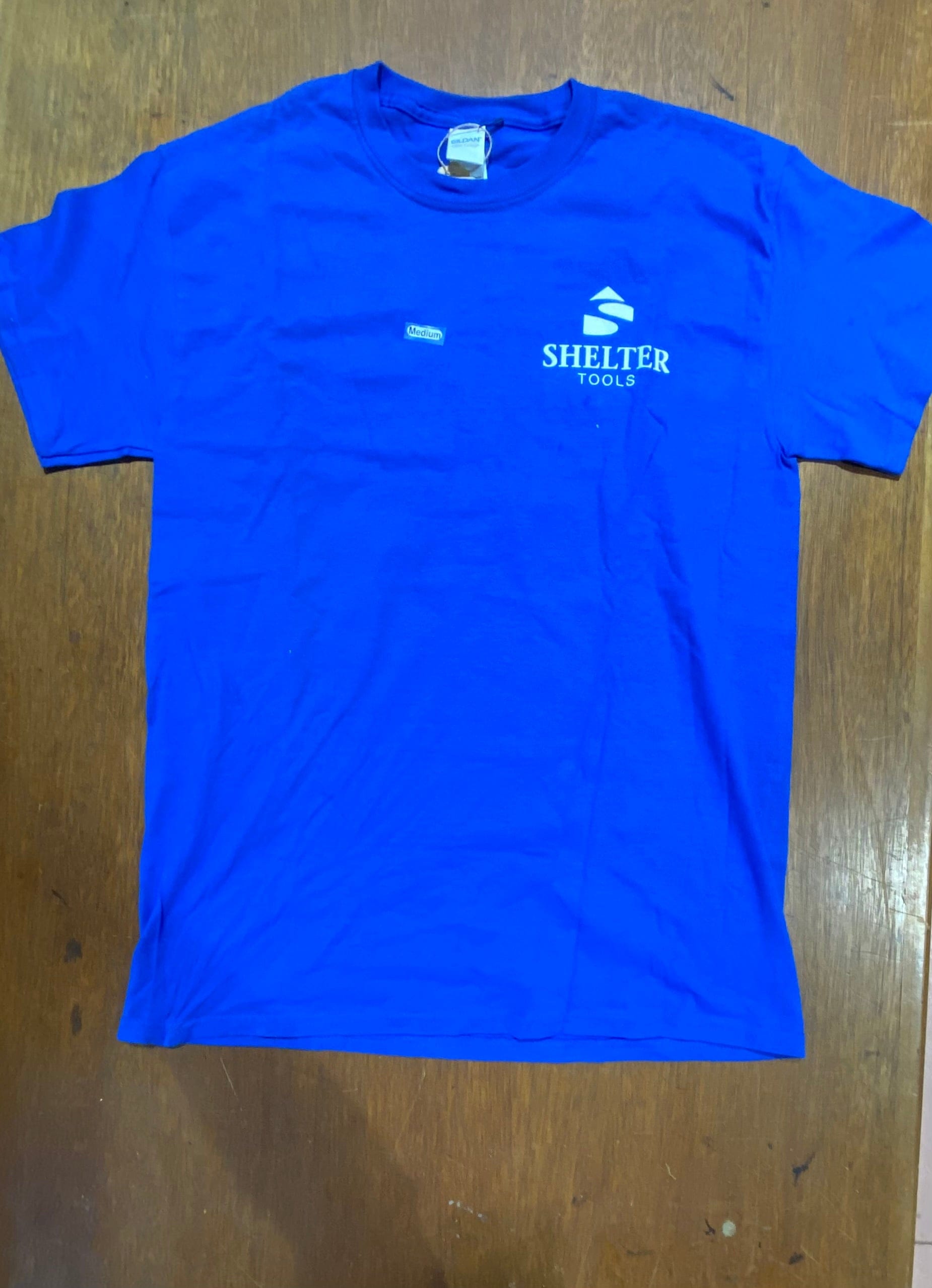 Shelter Tools T-Shirt by Shelter Institute, royal blue and made of 100% cotton, featuring the "Shelter Tools" logo and text screen printed on the left chest, displayed on a wooden surface.