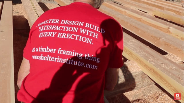 A person wearing a red Shelter Institute Satisfaction Tshirt with the text, "SHELTER DESIGN BUILD SATISFACTION WITH EVERY ERECTION. It's a timber framing thing. www.shelterinstitute.com," working with wooden beams.