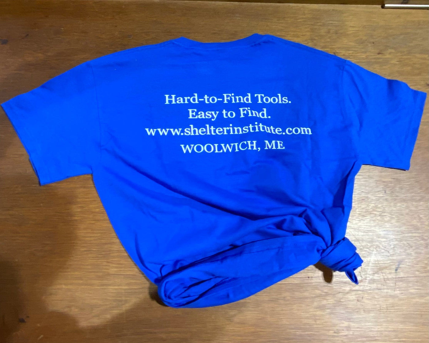 The Shelter Tools T-Shirt by Shelter Institute is royal blue, 100% cotton, with white Hard-to-Find Tools. Easy to Find. www.shelterinstitute.com WOOLWICH, ME text on the back; bottom tied in a knot.