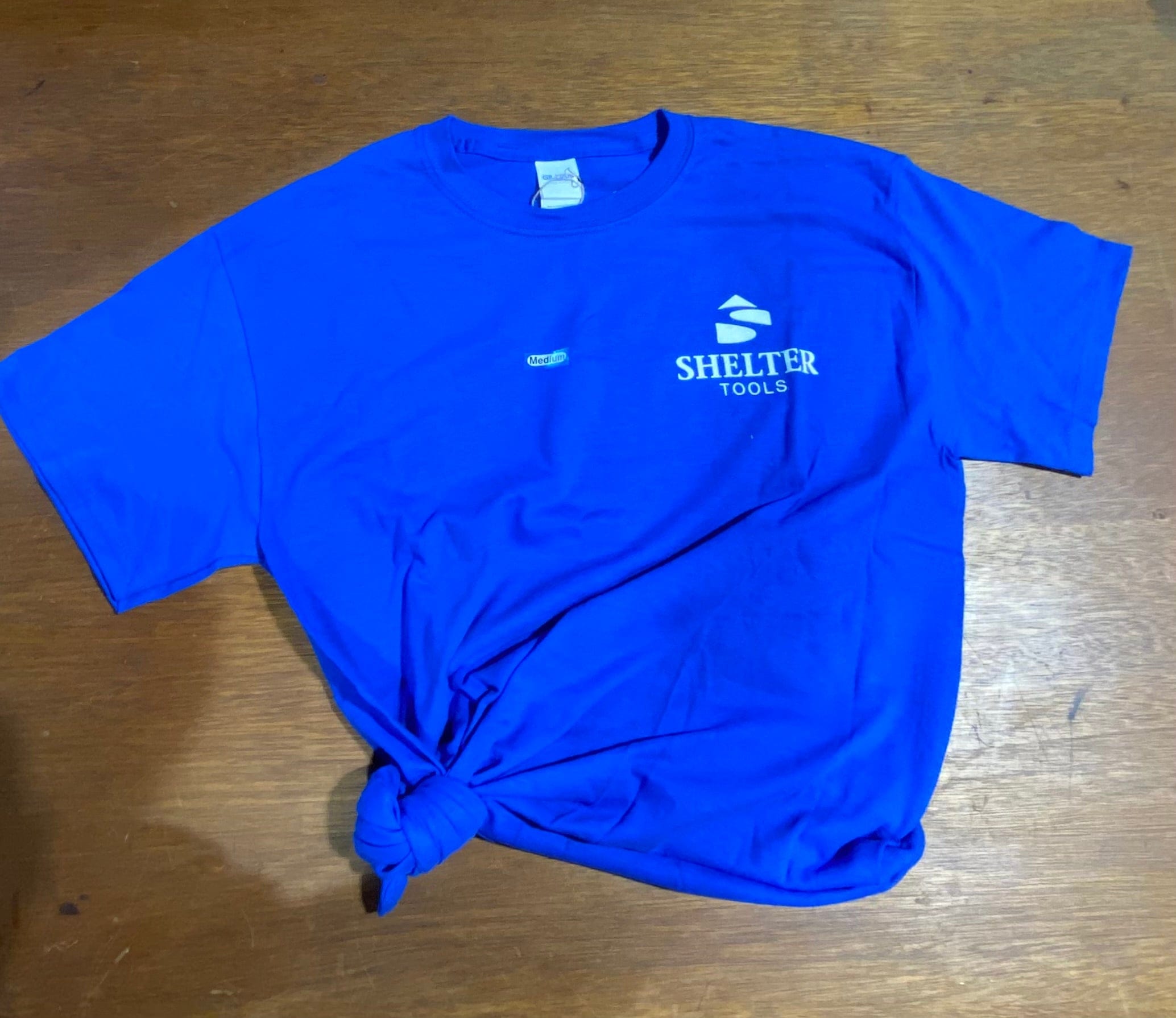 The Shelter Tools T-Shirt by Shelter Institute is royal blue, 100% cotton, with a bottom-left knot and the white Shelter Tools logo on the upper right chest, displayed flat on a wooden surface.